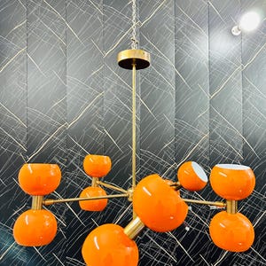 May include: A modern chandelier with eight orange globe-shaped lights. The fixture has a brass finish and is suspended from a chain. The lights are arranged on horizontal arms extending from a central stem. The background is a black wall with white lines.