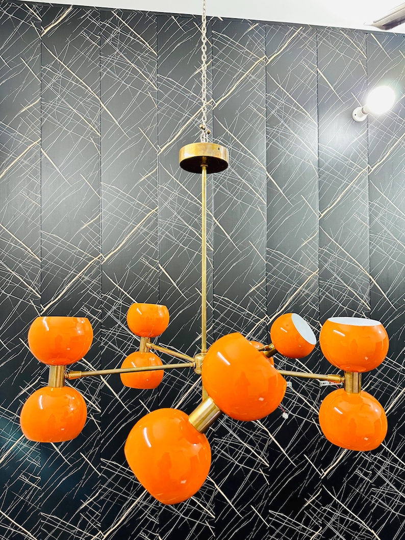 May include: A modern chandelier with eight orange globe-shaped lights. The fixture has a brass finish and is suspended from a chain. The lights are arranged on horizontal arms extending from a central stem. The background is a black wall with white lines.