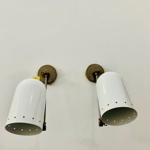 May include: A pair of white wall sconces with brass accents and a perforated design. The sconces are mounted on a white wall.