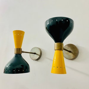 May include: Two wall sconces featuring a dual-cone design with glossy black and yellow finishes. Each lamp has a central brass-toned connector, a circular wall mount, and small perforated holes along the edges of the conical shades.