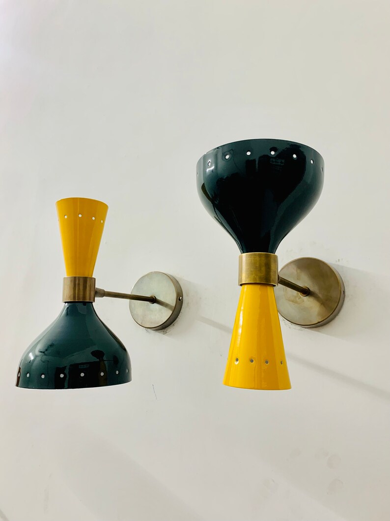 May include: Two wall sconces featuring a dual-cone design with glossy black and yellow finishes. Each lamp has a central brass-toned connector, a circular wall mount, and small perforated holes along the edges of the conical shades.