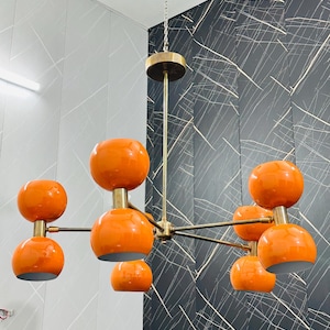 May include: A mid-century modern chandelier with eight orange globe shades. The fixture has a brass finish and hangs from a chain. The shades are a glossy orange color, and the fixture is suspended from the ceiling.