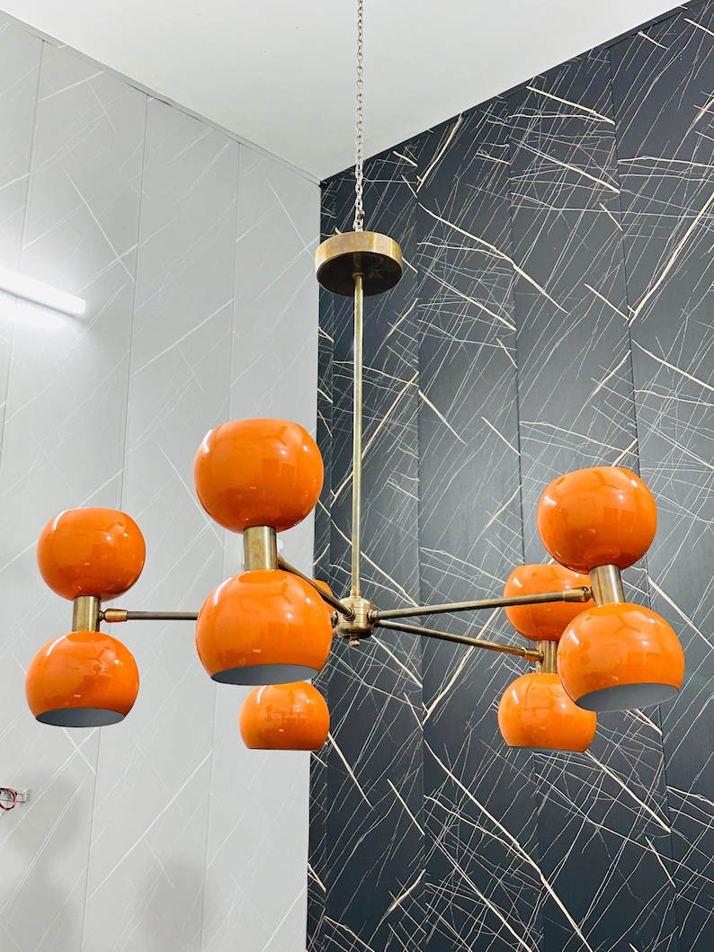 May include: A mid-century modern chandelier with eight orange globe shades. The fixture has a brass finish and hangs from a chain. The shades are a glossy orange color, and the fixture is suspended from the ceiling.