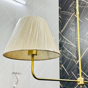 May include: A beige lampshade with a gold-colored metal frame. The lampshade is cone-shaped and has a textured surface. The lamp is hanging from the ceiling. The background is a light gray wall with a dark gray patterned wall.