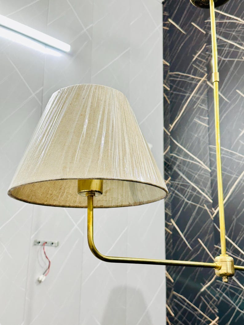 May include: A beige lampshade with a gold-colored metal frame. The lampshade is cone-shaped and has a textured surface. The lamp is hanging from the ceiling. The background is a light gray wall with a dark gray patterned wall.