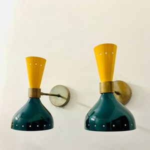 May include: Two wall sconces featuring a dual-tone design with a dark green bell-shaped base and a yellow conical top. Each lamp includes a brass-toned circular wall mount, a horizontal arm, and a central brass band. Small circular perforations are visible along the rims of both the yellow and green sections.