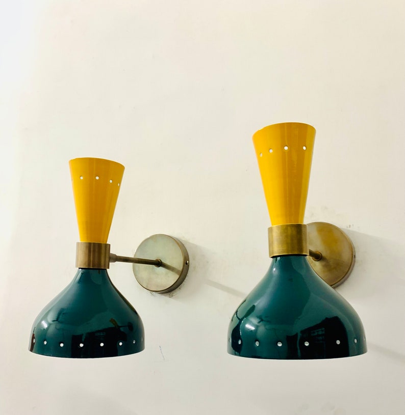 May include: Two wall sconces featuring a dual-tone design with a dark green bell-shaped base and a yellow conical top. Each lamp includes a brass-toned circular wall mount, a horizontal arm, and a central brass band. Small circular perforations are visible along the rims of both the yellow and green sections.