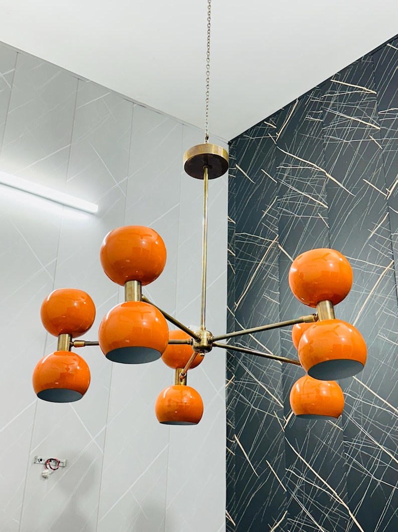 May include: A mid-century modern chandelier with eight orange globe shades. The fixture has a brass finish and hangs from a chain. The shades are a glossy orange and are evenly spaced around the central hub.