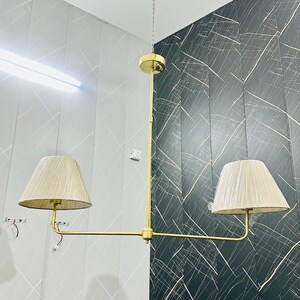 May include: A gold-toned, two-light pendant lamp with beige fabric shades. The lamp hangs from a chain and features a horizontal bar with two shades. The background shows a wall with a gray and black pattern.