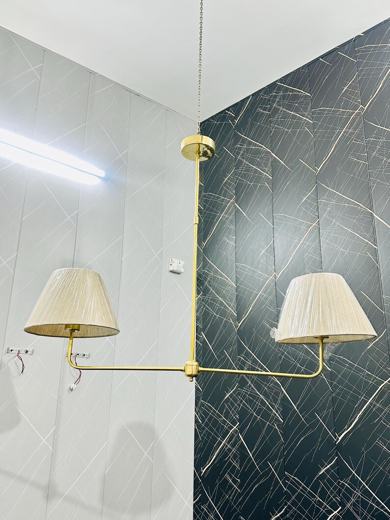 May include: A gold-toned, two-light pendant lamp with beige fabric shades. The lamp hangs from a chain and features a horizontal bar with two shades. The background shows a wall with a gray and black pattern.