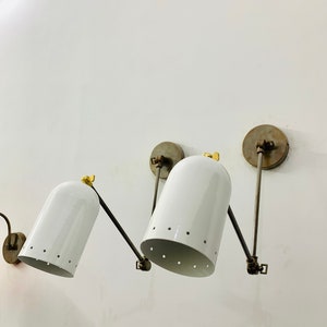 May include: Two white wall-mounted lights with adjustable arms and perforated shades. The lights have a brass finish and are mounted on a white wall.