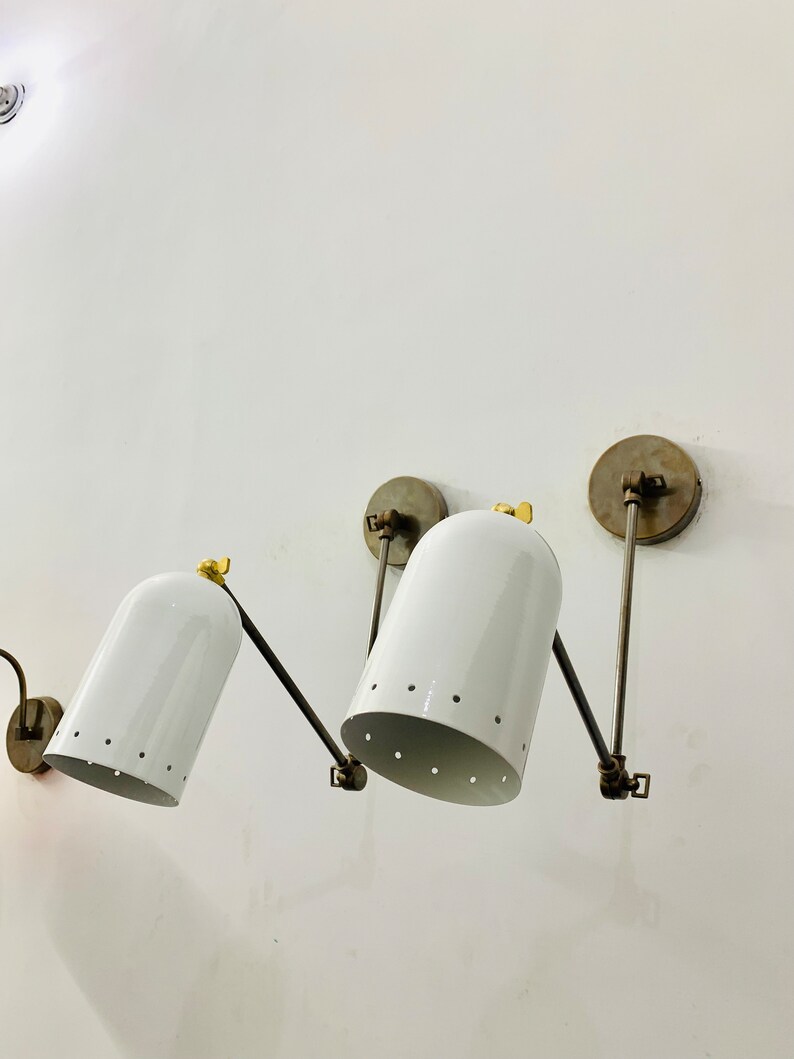 May include: Two white wall-mounted lights with adjustable arms and perforated shades. The lights have a brass finish and are mounted on a white wall.