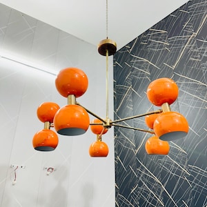 May include: A mid-century modern chandelier with eight orange globe shades. The fixture has a brass finish and is suspended from the ceiling by a chain. The shades are evenly spaced around the central hub.