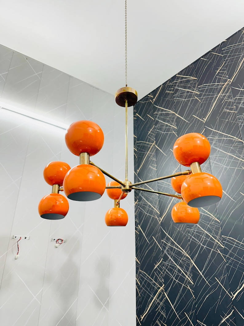May include: A mid-century modern chandelier with eight orange globe shades. The fixture has a brass finish and is suspended from the ceiling by a chain. The shades are evenly spaced around the central hub.