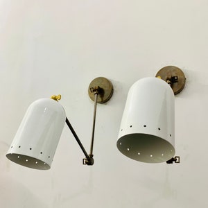 May include: Two white metal wall sconces with brass accents and a perforated design. The sconces are mounted on a white wall.