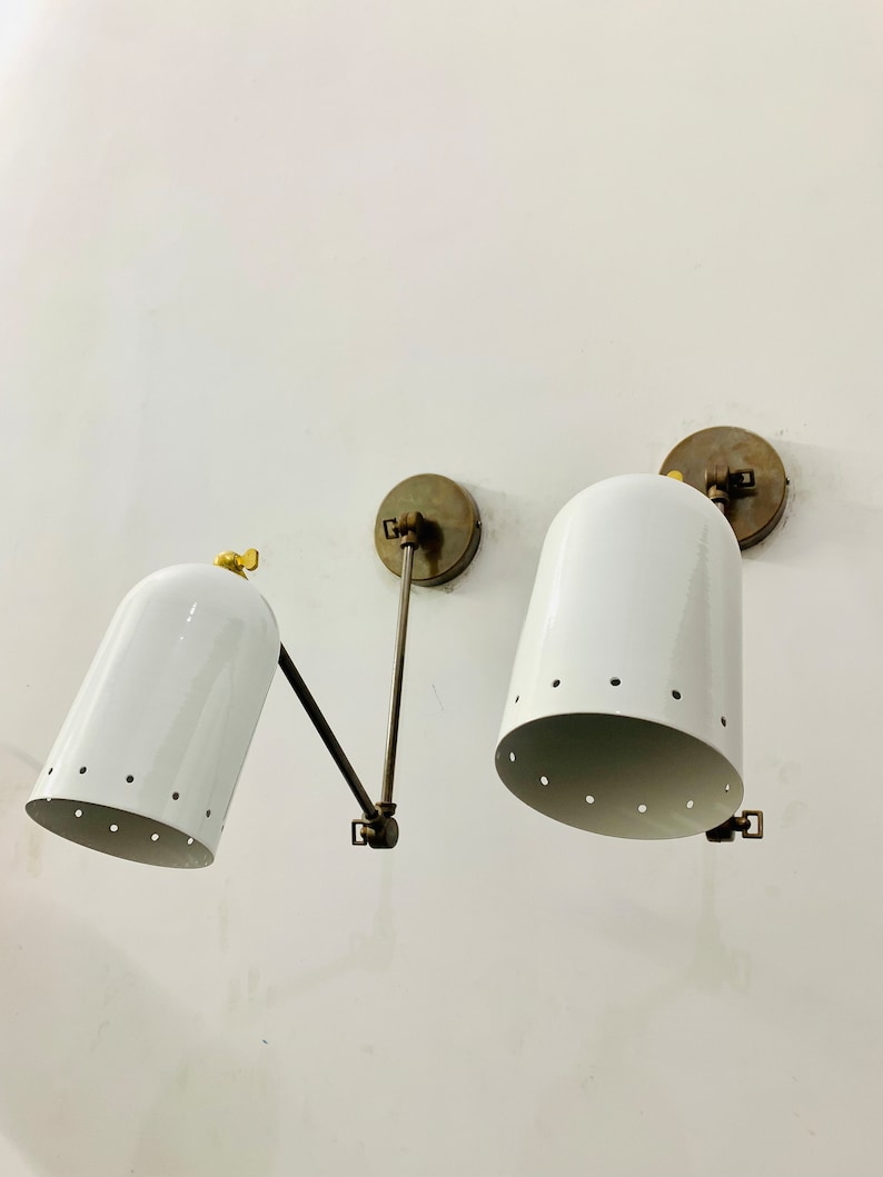 May include: Two white metal wall sconces with brass accents and a perforated design. The sconces are mounted on a white wall.