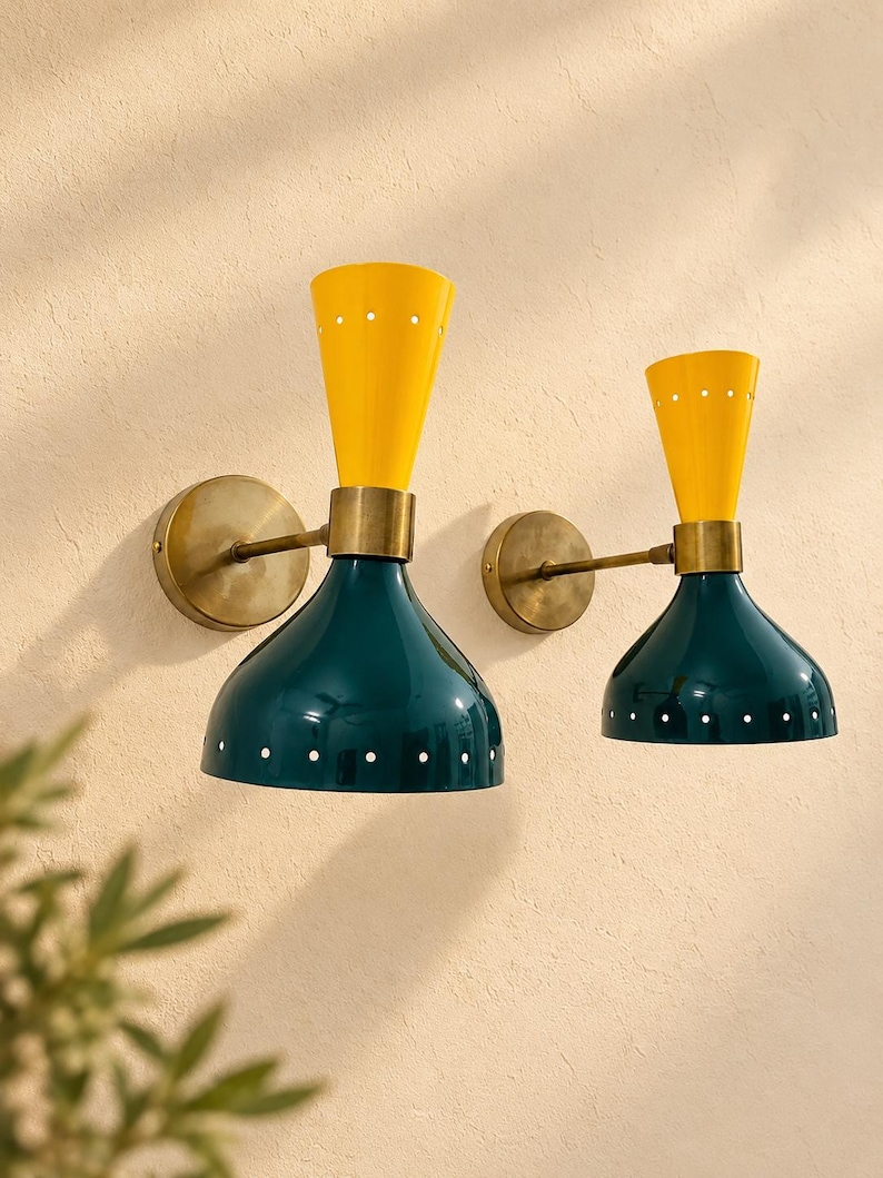 May include: Two wall-mounted lamps featuring a dual-cone design. Each lamp has a bright yellow upper cone and a dark teal lower cone, both accented with small white circular perforations. The fixtures are connected by brass-toned metal arms.