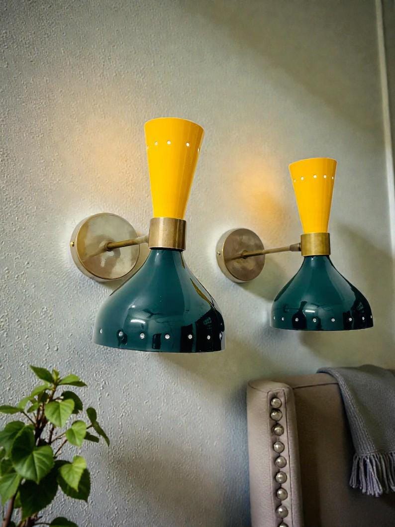 May include: Two wall-mounted lamps featuring a dual-tone design with a bright yellow conical top section and a dark teal bell-shaped bottom shade. The fixtures are connected by brass-toned arms to circular wall plates and include small white accents.