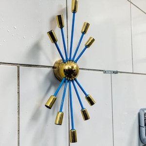 Puede incluir: Aplique de pared retro con centro de latón y accesorios de iluminación dorados. Los brazos son de color azul brillante. El aplique está fijado a una pared de azulejos blancos.