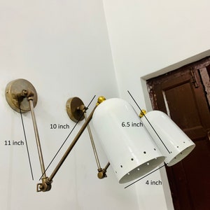 May include: A white wall-mounted light fixture with two adjustable arms. Each arm has a white, perforated shade with a gold-toned base. The fixture measures 11 inches long, 10 inches wide, 6.5 inches tall, and 4 inches deep.