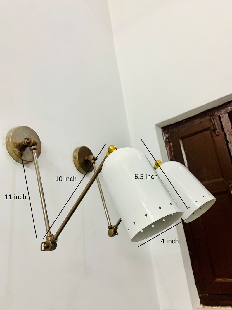 May include: A white wall-mounted light fixture with two adjustable arms. Each arm has a white, perforated shade with a gold-toned base. The fixture measures 11 inches long, 10 inches wide, 6.5 inches tall, and 4 inches deep.