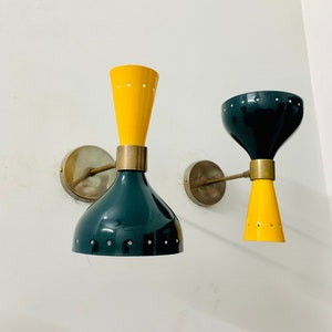 May include: Two wall sconces featuring dual-cone shades in yellow and dark green, connected by a central brass-toned metal band. Each fixture is mounted to the wall with a circular brass-toned plate and a slender horizontal arm.