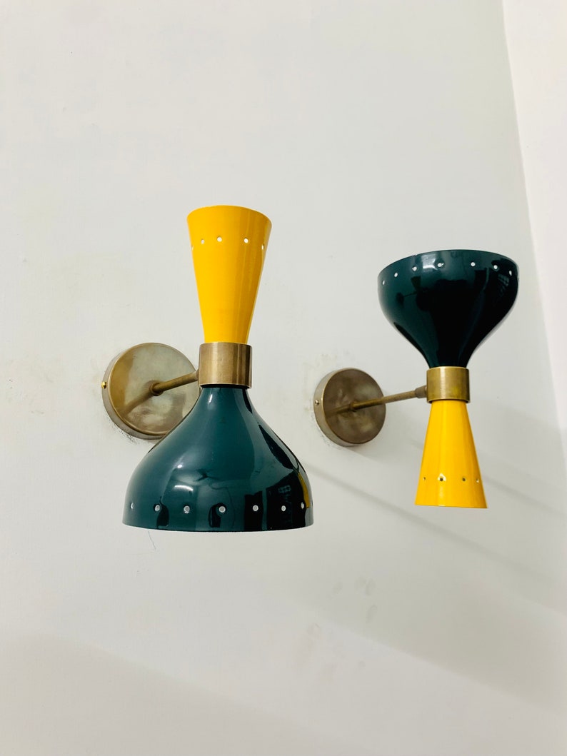 May include: Two wall sconces featuring dual-cone shades in yellow and dark green, connected by a central brass-toned metal band. Each fixture is mounted to the wall with a circular brass-toned plate and a slender horizontal arm.