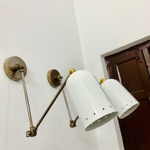 May include: A pair of white metal wall sconces with brass accents and adjustable arms. The sconces have a cylindrical shade with a row of small holes around the bottom.