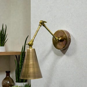 May include: A brass wall lamp featuring an adjustable arm and a conical shade. The lamp is fixed to a circular base and is constructed from a warm, metallic material. The design is minimalist and suitable for various interior styles.