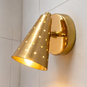 May include: A wall-mounted light fixture featuring a conical brass shade with a perforated pattern. The shade is attached to a circular brass base plate by a small adjustable arm. A warm light glows from within the cone, illuminating the holes.