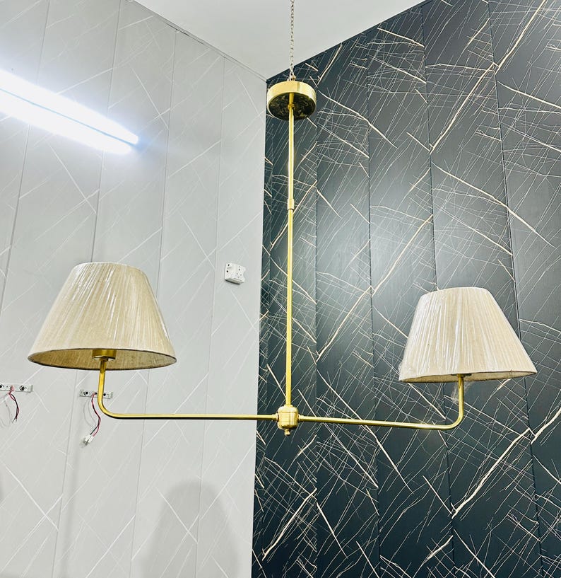 May include: A gold-toned, two-light chandelier with beige fabric shades. The fixture has a central vertical rod and a horizontal arm extending to each shade. The shades are cone-shaped.