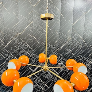 May include: A mid-century modern chandelier with a brass finish and ten orange globe shades with white interiors. The fixture hangs from a chain and a brass ceiling mount. The design is symmetrical and eye-catching.