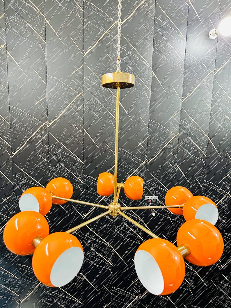May include: A mid-century modern chandelier with a brass finish and ten orange globe shades with white interiors. The fixture hangs from a chain and a brass ceiling mount. The design is symmetrical and eye-catching.