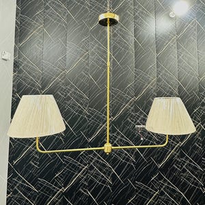 May include: A gold-toned, two-light pendant lamp with beige fabric shades. The lamp hangs from a chain and features a horizontal bar with two shades. The background is a black wall with white linear patterns.