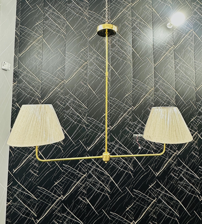May include: A gold-toned, two-light pendant lamp with beige fabric shades. The lamp hangs from a chain and features a horizontal bar with two shades. The background is a black wall with white linear patterns.