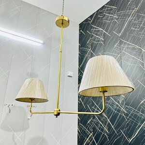 May include: A brass chandelier with two beige fabric lampshades. The fixture hangs from a chain and features a central vertical rod with horizontal arms supporting the shades. The lampshades are cone-shaped and the fixture has a modern design.