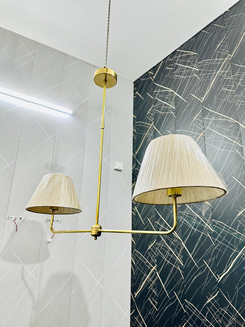 May include: A brass chandelier with two beige fabric lampshades. The fixture hangs from a chain and features a central vertical rod with horizontal arms supporting the shades. The lampshades are cone-shaped and the fixture has a modern design.
