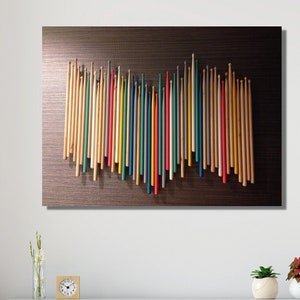 Pode incluir: Uma peça de arte de parede com uma coleção de baquetas de madeira coloridas dispostas em forma de onda sobre um fundo marrom escuro.