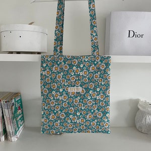 Tote bag premium Marguerites 

Le fond du tote bag est Bleu turquoise foncé. Les marguerites ont des pétales blanches et un centre jaune pollen. 
Le motif est assez chargé : les marguerites recouvrent tout le tote bag même les hanses.