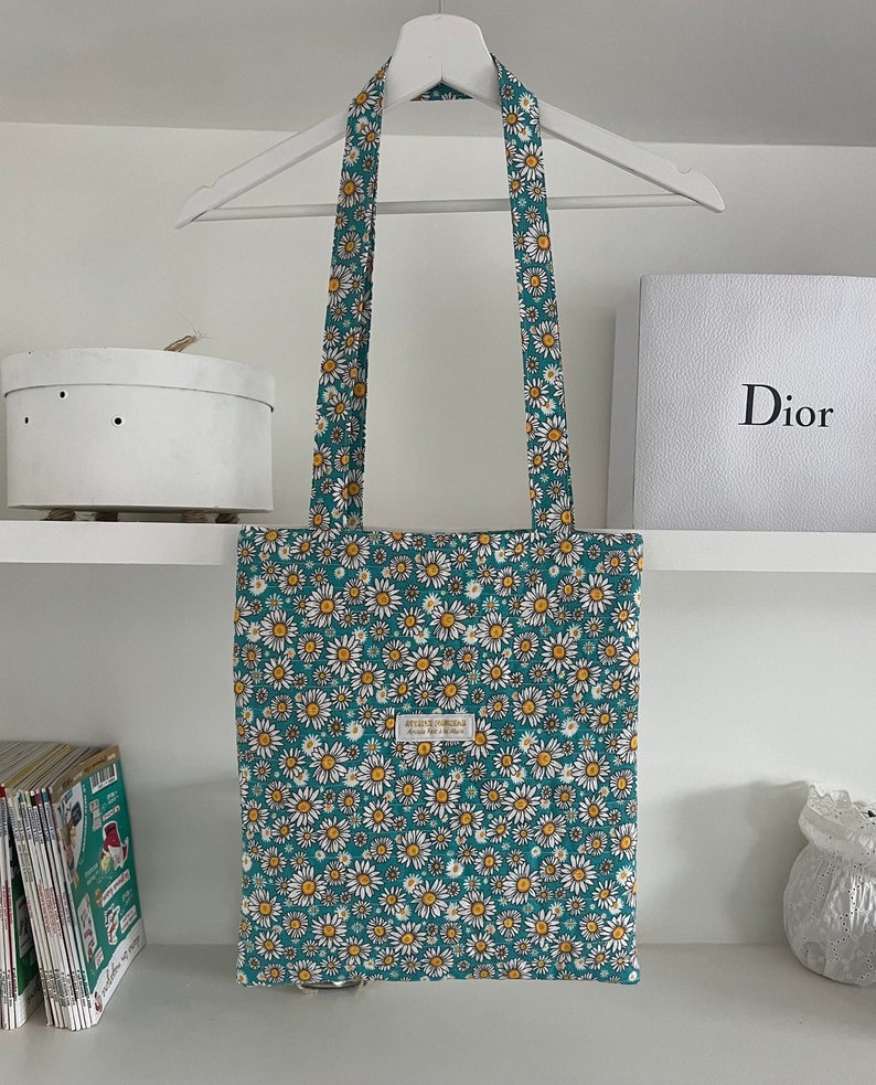 Tote bag premium Marguerites 

Le fond du tote bag est Bleu turquoise foncé. Les marguerites ont des pétales blanches et un centre jaune pollen. 
Le motif est assez chargé : les marguerites recouvrent tout le tote bag même les hanses.