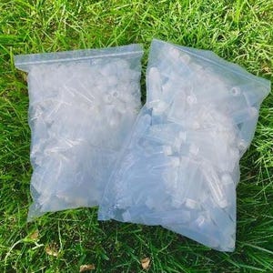 May include: Two clear plastic bags filled with numerous small, transparent vials with white caps. The bags are sealed and rest on a bed of green grass, suggesting an outdoor setting.