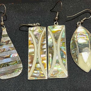 May include: A collection of five pairs of silver earrings with iridescent abalone shell inlays. The earrings feature various shapes, including teardrop, rectangular, and oval designs. Each earring has a silver hook for pierced ears.
