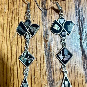May include: A pair of drop earrings featuring silver-tone metal frames with black inlay and iridescent shell fragments. Each earring consists of three connected geometric shapes: a clover-like top, a diamond center, and a teardrop bottom.
