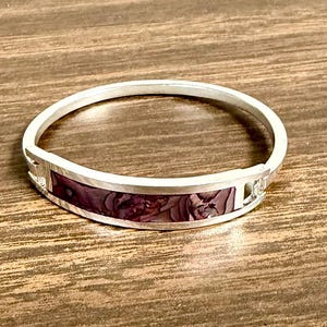May include: A silver-toned bracelet with a rectangular, dark purple inlay featuring a floral pattern. The bracelet has a hinged design and is set against a wooden surface.