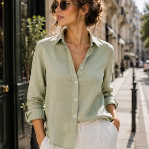 May include: A light sage green long-sleeved button-down shirt with a classic collar and white buttons, paired with white trousers. The shirt features a textured fabric finish, long sleeves with rolled cuffs, and a relaxed fit with a straight hemline.