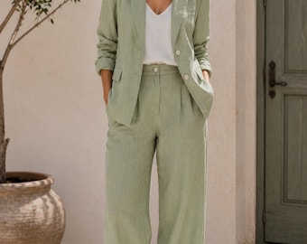 May include: A sage green two-piece suit consisting of a single-breasted blazer with notched lapels and two buttons, paired with high-waisted wide-leg trousers featuring a front button closure and side pockets, worn over a simple white V-neck top.