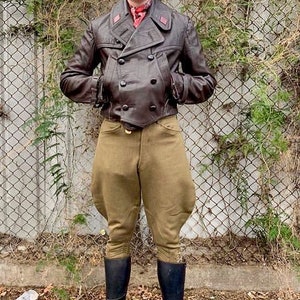 Puede incluir: Un hombre con una chaqueta de cuero marrón, una gorra a cuadros, botas de montar marrones y pantalones de montar de color caqui. Está de pie frente a una valla de alambre.