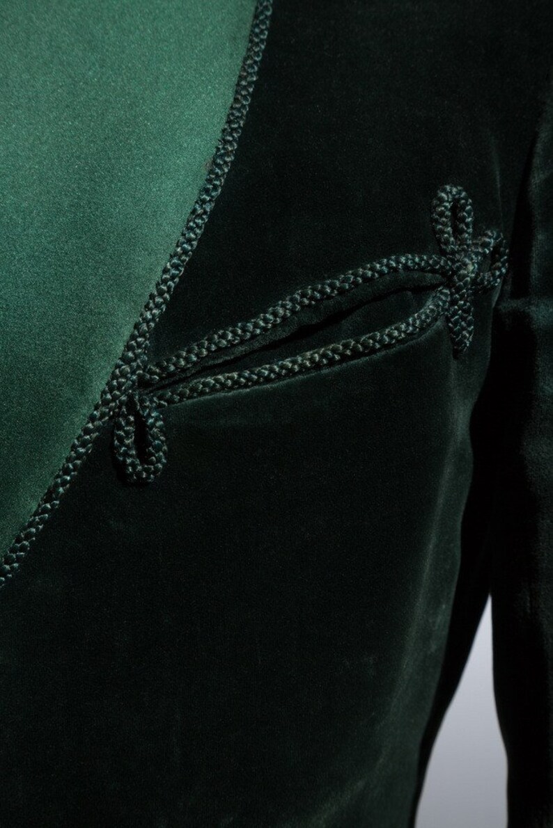 Pu&ograve; includere: Una giacca di velluto verde scuro con un bordo decorativo in corda nera. La giacca ha un dettaglio di tasca unico con un bordo in corda.