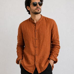 May include: A long-sleeved, burnt orange shirt with a mandarin collar and a button-down front. The shirt features a textured fabric finish, long sleeves with cuffs, and a straight hemline. The garment is paired with plain black trousers.