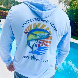 May include: A light blue long-sleeved hooded shirt with a graphic of a fish jumping out of the water with a sun and flags behind it. The text "UXEM FISHING TEAM" is above the fish and "MIAMI BAHAMAS" is below the fish.